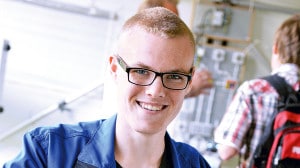 Ein bebrillter junger Mann im blauen Hemd und kurzen Haaren lächelt in die Kamera.