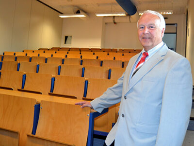 Ein Mann in grauen Anzug und mit roter Krawatte steht in einem Hörsaal.
