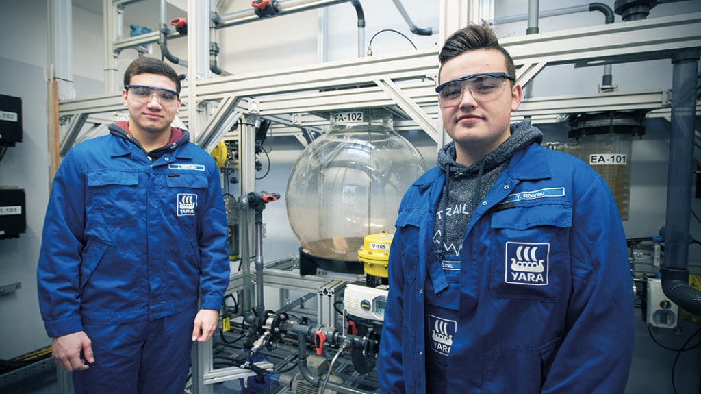 Zwei junge Männer in blauer Arbeitskleidung und mit Schutzbrille blicken in die Kamera.