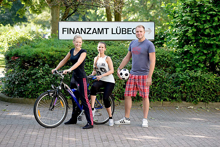 Drei junge Leute in Sportkleidung vor einem Schild mit der Aufschrift „Finanzamt Lübeck."