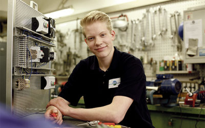 Ein blonder junger Mann im dunklen Shirt sitzt in einer Werkstatt und lächelt in die Kamera.