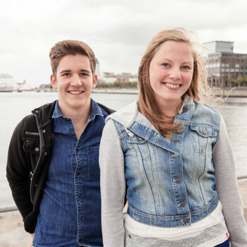 Zwei junge Menschen stehen vor einem Gewässer und lächeln in die Kamera.