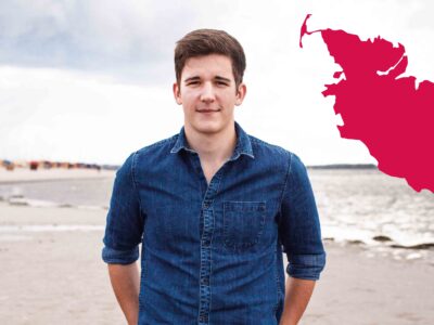 Ein junger Mann blickt am Strand in die Kamera.