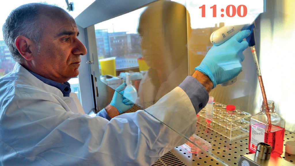 Ein Mann im weißen Kittel steht in einem Labor. Er trägt Handschuhe und taucht eine dicke Kanüle in eine rote Flüssigkeit. 