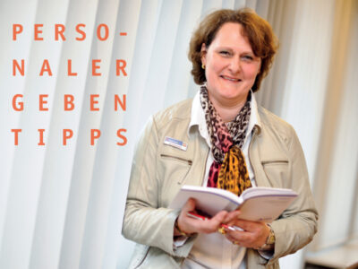 Eine Frau miFrau Reimers gibt Bewerbungstippsttleren Alters hält ein aufgeschlagenes Buch in den Händen und lächelt in die Kamera.