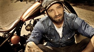 Ein Mann mit Bart, Mütze und Jeanshemd sitzt vor einem Motorrad.
