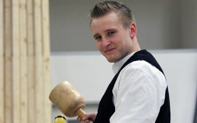 Ein blonder junger Mann steht mit einem Holzhammer vor einer Holzwand.