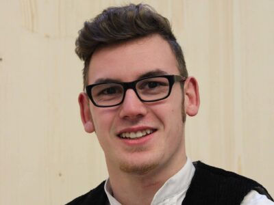 Ein junger Mann mit Brille steht vor einer Holzwand