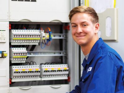 Ein blonder junger Mann im blauen Overall sitzt vor einem technischen Gerät.