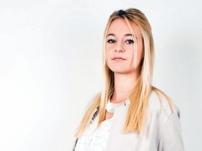 Eine blonde junge Frau mit hellem Oberteil vor einer grauen Wand.
