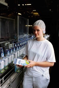 Eine junge Frau in weißer Arbeitskleidung steht vor einem Fließband mit Milchkartons.