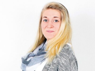 Eine junge blonde Frau vor einer grauen Wand schaut in die kamera.