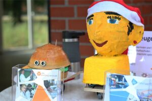 Zwei selbstgebaute Miniroboter sind mit Pappmaché verkleidet und auf einem Tisch ausgestellt. Einer sieht aus wie ein Hamburger mit Augen, der andere ähnelt einer gelb bemalten Büste mit einer Weihnachtsmannmütze.