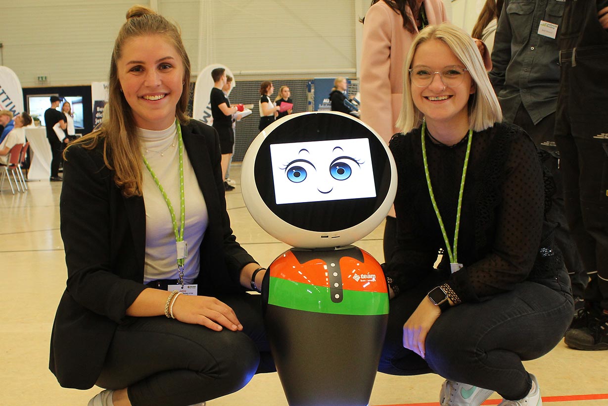 Zwei blonde junge Frauen hocken neben einem Roboter, dessen Gesicht aus einem Bildschirm erscheint.