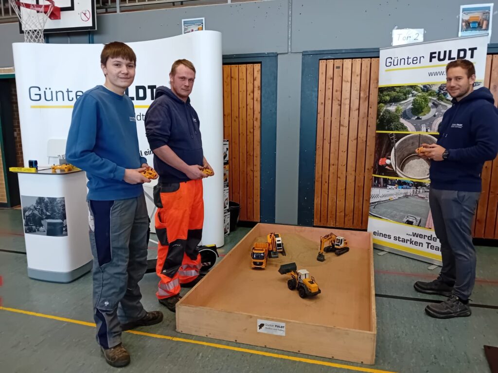 Das macht auch den Azubis Devin und Yanik sowie Straßenbaumeister und Ausbildungsleiter Yanik Pehrs Spaß (v. l. n. r.): Am Stand der Günter FULDT Straßen- u. Tiefbau GmbH konnten die Schülerinnen und Schüler mit ferngesteuerten Baggern fahren